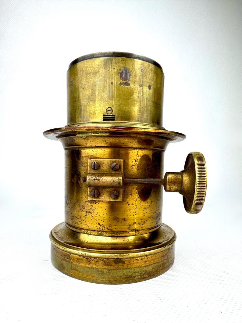 May include: Antique brass lamp component with a cylindrical body, a circular base, and a ridged knob. The brass has a tarnished, aged appearance, suggesting its vintage. The component features a series of screws and a small lever. The overall design is industrial.