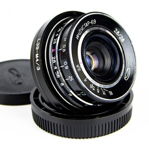 May include: A black camera lens with a silver ring and markings. The lens has a black cap and the text "L39-M4/3" and "ИНДУСТАР-69" is visible on the lens.