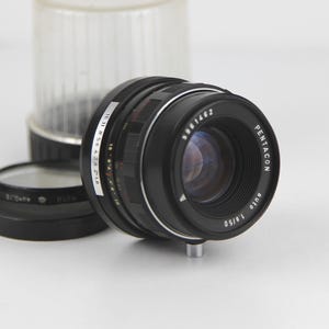 May include: A black Pentacon Auto 1:1.5/50 lens with a silver ring and markings. The lens has a black lens cap and a silver aperture ring.
