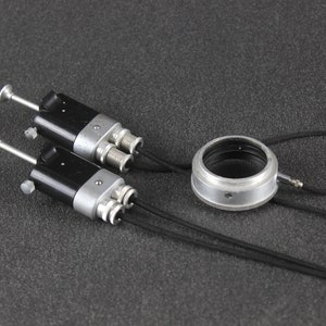 Può includere: Due accessori per fotocamera neri e argentati con cavi neri. Gli accessori hanno una manopola argentata e un connettore argentato con due spine. È anche raffigurato un anello argentato con un elastico nero.