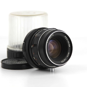 May include: A black Pentacon Auto 1.8/50 lens with a silver ring around the lens barrel. The lens is attached to a black lens cap.