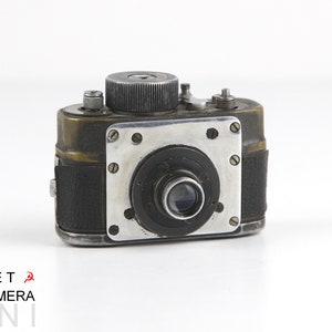 May include: A vintage, black and brass spy camera with a silver lens. The camera has a textured black leather covering and a silver metal plate with screws around the lens.