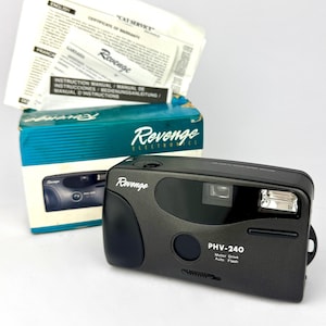 以下が含まれることがあります： 黒色のRevenge PHV-240フィルムカメラ。内蔵フラッシュと「PHV-240 Motor Drive Auto Flash」の文字があります。カメラはオリジナルの箱と取扱説明書と一緒に写っています。