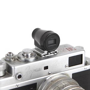 Puede incluir: Una cámara de película de 35 mm plateada y negra con un fotómetro incorporado. La cámara tiene una cubierta de imitación de cuero negro y un cuerpo de metal plateado. La cámara tiene un visor de telémetro y una lente con una tapa de lente negra. La cámara tiene una correa de cuero negra.