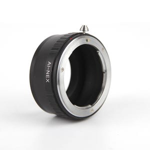 Op de afbeelding: Zwarte AI-NEX lensadapter met een zilverkleurige ring en een kleine zilveren knop. Ontworpen om AI-vatting lenzen aan Sony E-mount camera's te koppelen. De adapter wordt getoond tegen een witte achtergrond.