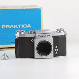 Puede incluir: Una cámara Praktica plateada y negra con una caja azul. La cámara tiene una lente plateada y un cuerpo negro.