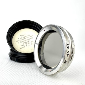 Può includere: Un filtro per obiettivo argentato con custodia protettiva nera. Il filtro ha un bordo scanalato con segni e un centro in vetro trasparente. La custodia è aperta, rivelando testo in cirillico. Il filtro è per una fotocamera Kiev.