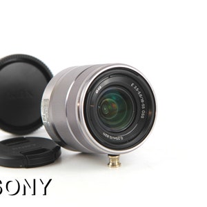 Puede incluir: Un objetivo Sony E 3.5-5.6/18-55 OSS plateado con una tapa de lente negra y una parasol negra.