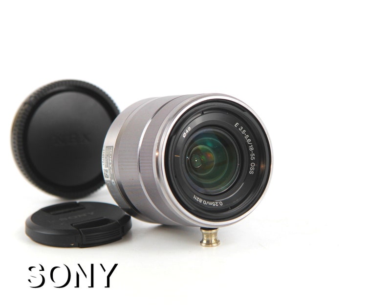 Puede incluir: Un objetivo Sony E 3.5-5.6/18-55 OSS plateado con una tapa de lente negra y una parasol negra.