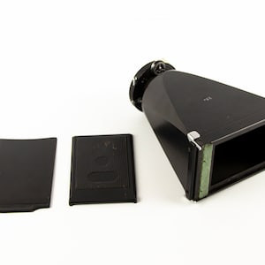 Puede incluir: Un fuelle de cámara negro con dos placas negras separadas. El fuelle tiene una abertura rectangular con acolchado verde. El accesorio de la cámara está sobre un fondo blanco.