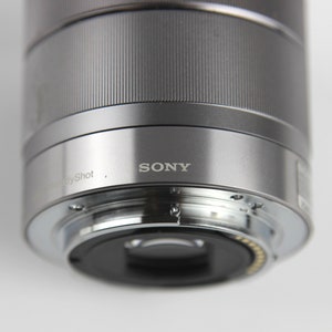 Puede incluir: Un objetivo de c&aacute;mara plateado con la palabra "SONY" impresa en &eacute;l. El objetivo est&aacute; conectado a un cuerpo de c&aacute;mara.