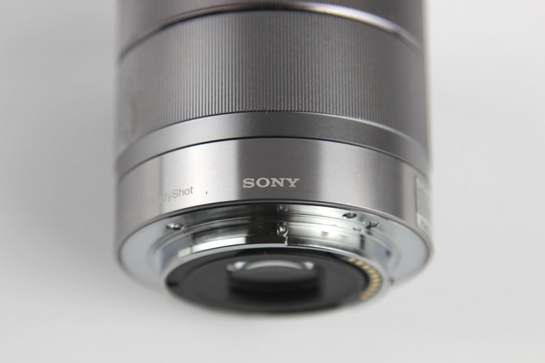 Puede incluir: Un objetivo de c&aacute;mara plateado con la palabra "SONY" impresa en &eacute;l. El objetivo est&aacute; conectado a un cuerpo de c&aacute;mara.