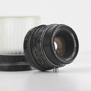 May include: A black Pentacon 135/3.5 lens with a silver ring around the lens barrel. The lens is in focus and the aperture is open.