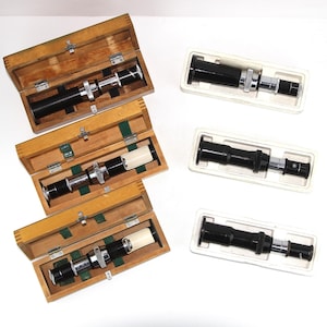 May include: Four microscope objectives in wooden and plastic cases. The objectives are black with silver accents and have a white ring around the lens.