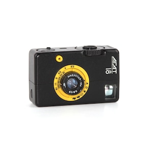 Op de afbeelding: Een zwarte en gele 35mm filmcamera met de tekst "AGAT 18" aan de zijkant. De camera heeft een gele lens met de tekst "INDUSTAR-104" en "2.8/28" erop gedrukt.
