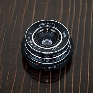 May include: A black and silver camera lens with the text "ИНДУСТАР-69" and "28/28" printed on the side. The lens has a silver ring with numbers and markings for aperture settings.