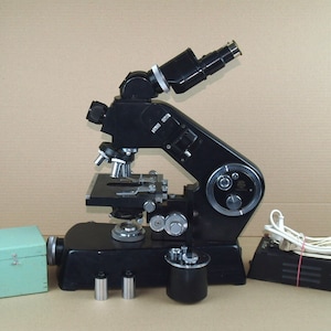 Puede incluir: Un microscopio negro con una fuente de luz y una platina para observar láminas. El microscopio tiene una base negra y una perilla plateada para ajustar el enfoque. Hay tres pequeños recipientes cilíndricos y una caja de madera con una tapa con bisagras.