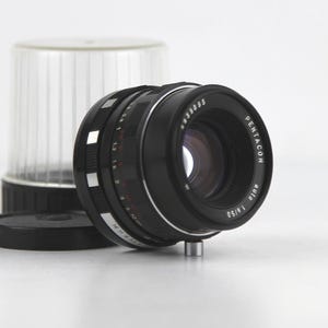 May include: A black Pentacon Auto 1.8/50 camera lens with a clear plastic flash attachment and a black lens cap. The lens has white markings and a silver base. The lens is on a white surface.