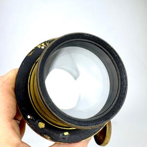 May include: A vintage camera lens with a large, clear glass element. The lens has a black and gold metal casing with a ribbed texture and a brass adjustment knob. The lens is held by a hand against a white background.