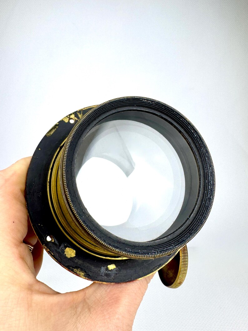 May include: A vintage camera lens with a large, clear glass element. The lens has a black and gold metal casing with a ribbed texture and a brass adjustment knob. The lens is held by a hand against a white background.