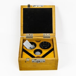 Puede incluir: Una caja de madera con tapa con bisagras, que contiene tres objetos circulares negros y un pequeño trozo de papel blanco. La caja está abierta y el interior está forrado con fieltro negro.