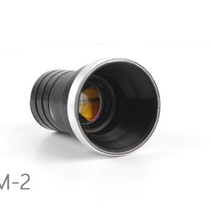 Puede incluir: Un objetivo de cámara negro y plateado con un anillo plateado alrededor del frente. El objetivo tiene una tapa de lente de goma negra.