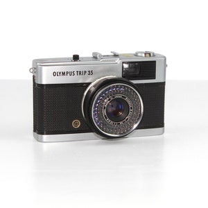 Op de afbeelding: Een zilveren Olympus Trip 35 filmcamera met een zwart, getextureerd lichaam en een zilveren lens met een blauwe ring rond de rand.