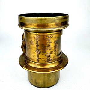 May include: Antique brass camera lens with the text "Portrait Objectiv" and "Rodens & Co. M&uuml;nchen". The lens has a cylindrical body with a flared base and top. The brass has a worn, aged appearance.