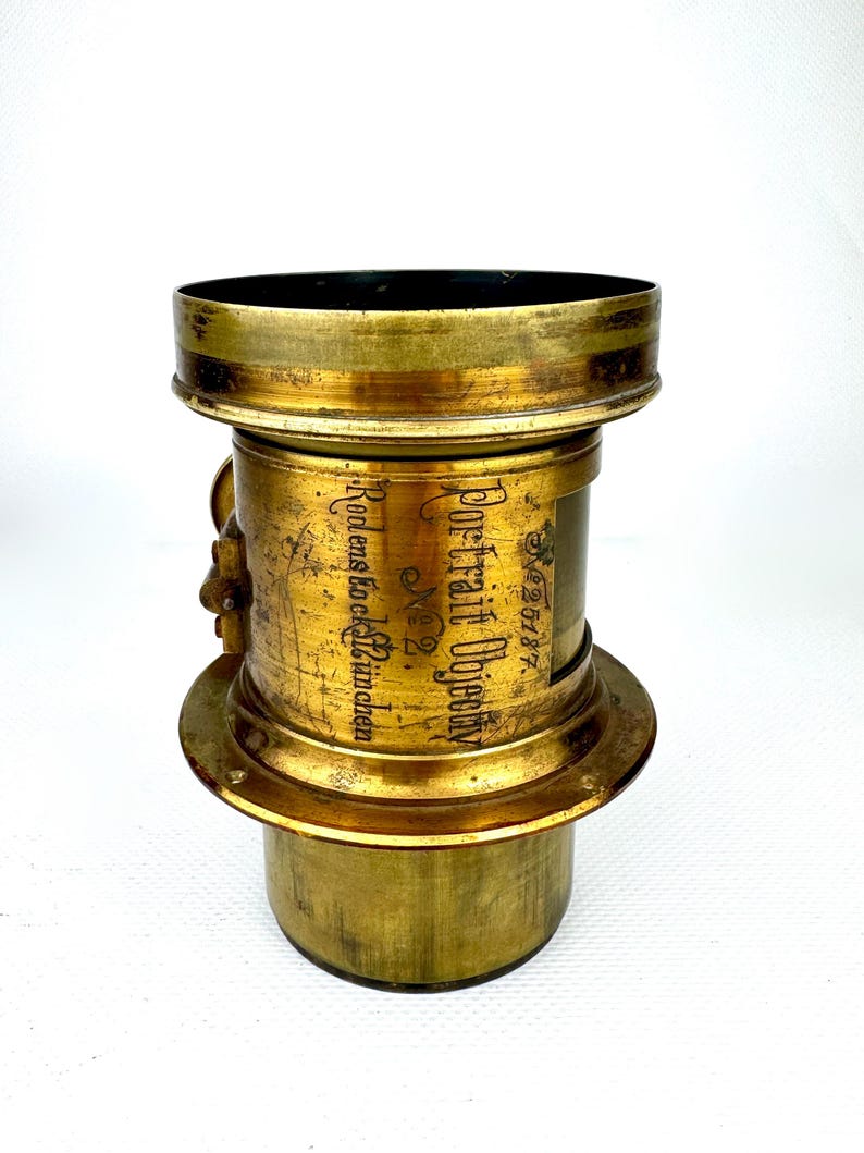 May include: Antique brass camera lens with the text "Portrait Objectiv" and "Rodens & Co. M&uuml;nchen". The lens has a cylindrical body with a flared base and top. The brass has a worn, aged appearance.