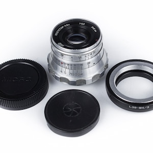 May include: A silver camera lens with black lens caps and a black adapter ring. The lens has the text "INDUSTAR-26u" and "1:2.8" printed on it. The adapter ring has the text "L39-M4/3" printed on it.