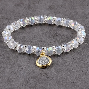 May include: A clear crystal beaded bracelet with silver accents and a gold charm. The charm is round with a detailed design. The bracelet is on a gray surface.