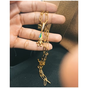 May include: A gold-toned chain necklace featuring a pendant of a small figure wearing a pointed hat with rainbow-colored stripes. The pendant is attached to the chain by a small, circular bail encrusted with clear, sparkling stones.