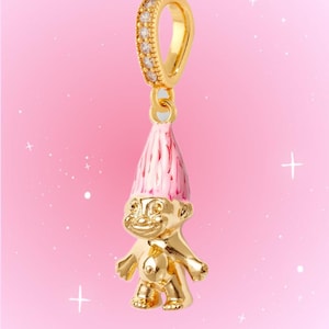 May include: A gold-toned troll charm featuring a shiny metallic body and a tall, pointed pink enamel hat. The charm is suspended from a gold-toned bail that is encrusted with small, clear, sparkling stones, designed for attachment to a bracelet or necklace.