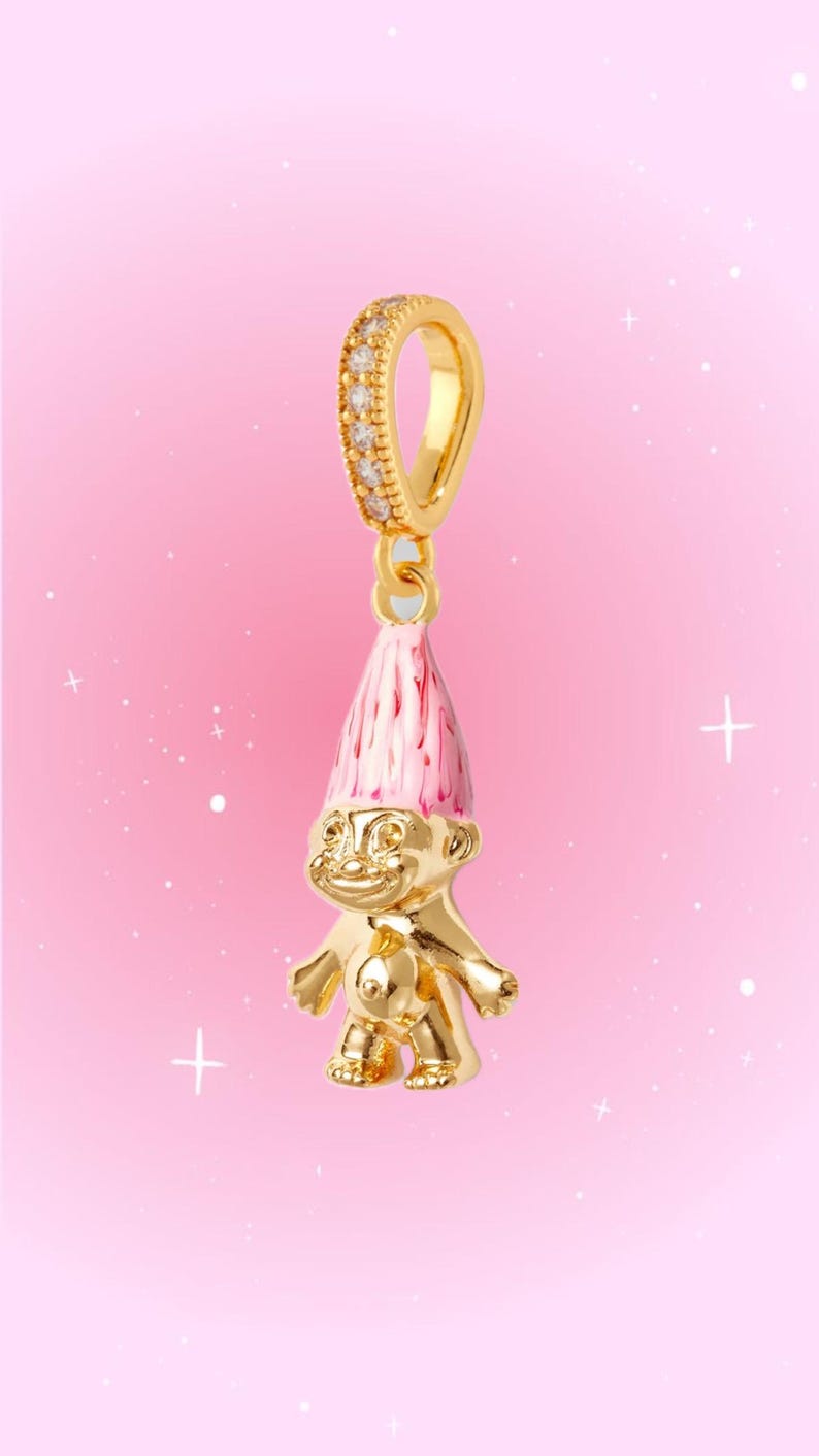 May include: A gold-toned troll charm featuring a shiny metallic body and a tall, pointed pink enamel hat. The charm is suspended from a gold-toned bail that is encrusted with small, clear, sparkling stones, designed for attachment to a bracelet or necklace.