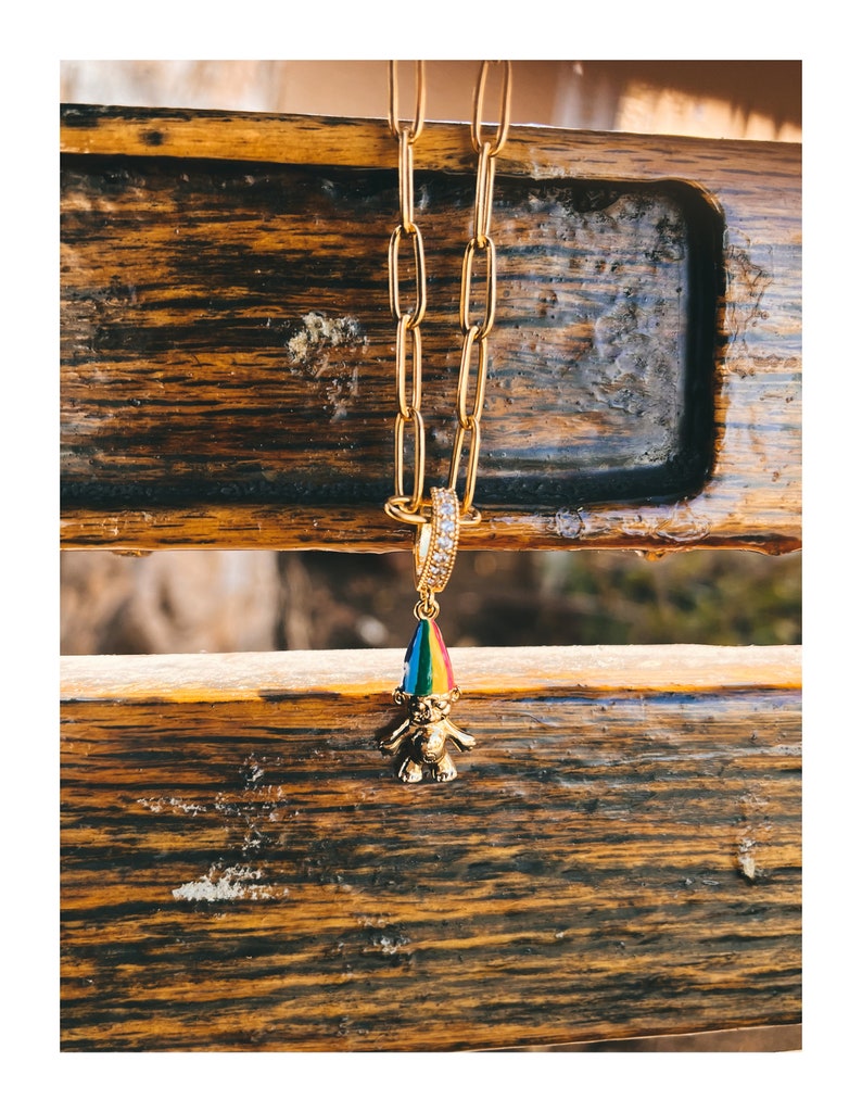 Puede incluir: Un collar de cadena de oro con un pequeño amuleto de gnomo colorido. El gnomo lleva un gorro de arcoíris y tiene un cuerpo dorado.