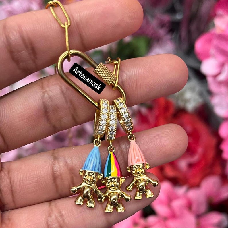 May include: A gold-toned carabiner-style clasp keychain featuring three small gold-toned troll charms with colorful enamel hats. The charms hang from individual gold-toned hoops encrusted with clear crystals. The text Artesaniask is visible.