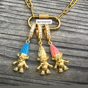 May include: A gold-toned necklace featuring a carabiner-style clasp with three dangling troll charms. Each charm has a gold body and a colored hat in blue, gold, or pink. The carabiner is accented with small clear stones. Text reads @artesaniask.