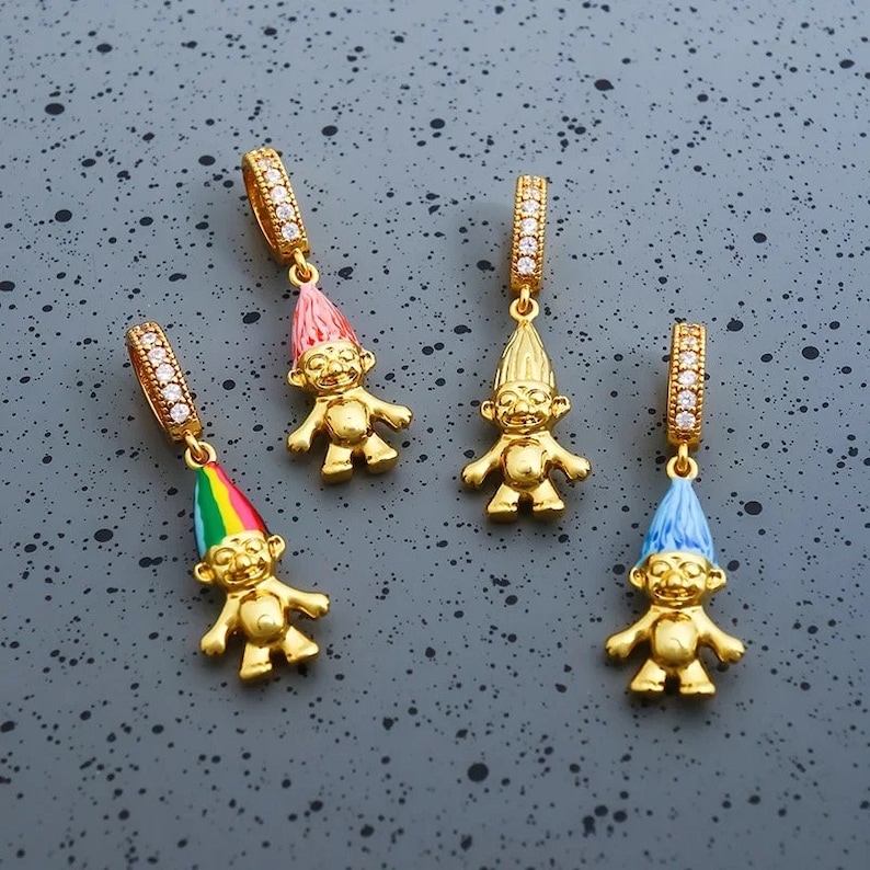May include: Four gold-toned troll charms, each featuring a small bail encrusted with clear crystals. Three trolls have colorful enamel hats in pink, rainbow, and blue, while one troll has a solid gold-toned hat. Each figure has a distinct facial expression.