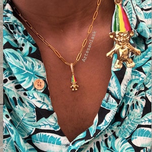 May include: A gold paperclip chain necklace featuring a small troll pendant with a multicolored striped hat and a matching large troll earring with a multicolored striped hat, worn with a teal and black tropical leaf print button-down shirt.