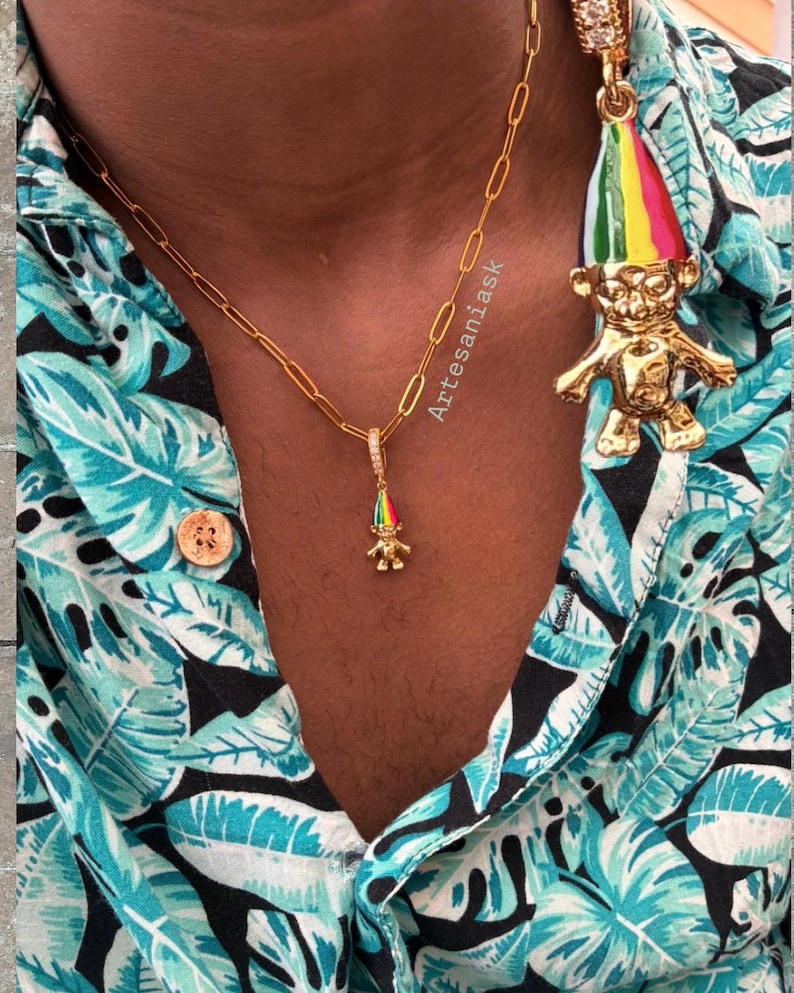May include: A gold paperclip chain necklace featuring a small troll pendant with a multicolored striped hat and a matching large troll earring with a multicolored striped hat, worn with a teal and black tropical leaf print button-down shirt.