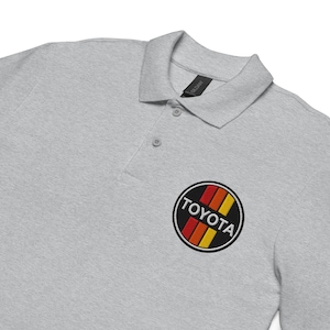 May include: A light gray polo shirt featuring a classic collar, a two-button placket, and an embroidered circular patch on the chest. The patch displays the word TOYOTA in white capital letters over three vertical stripes in red, orange, and yellow.