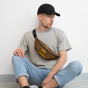 Könnte beinhalten: Braune Bauchtasche mit dem gelben Schriftzug "UPS STRONG". Die Bauchtasche hat einen schwarzen, verstellbaren Riemen und eine Schnalle. Die Person im Bild trägt eine schwarze Baseballkappe, ein graues T-Shirt und blaue Jeans.