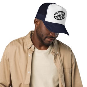 May include: A navy blue and white trucker hat featuring a white front panel with a black graphic of a coffee bean shape containing the text INSERT COFFEE TO BEGIN. The cap has a dark navy blue brim and a mesh back panel for ventilation.