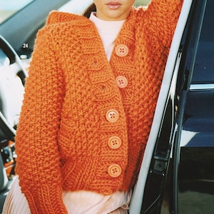 May include: A bright orange knitted cardigan with a button closure. The cardigan has a textured knit pattern and is worn by a person with brown hair and bangs.