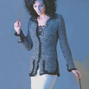 May include: A gray and black tweed jacket with black fringe trim and buttons. The jacket has two front pockets and is paired with a white skirt. The model is holding a black accessory.
