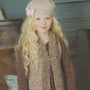 May include: A young girl wearing a brown and beige knitted vest with a brown polka dot dress and a beige knitted hat with a pink butterfly accent.