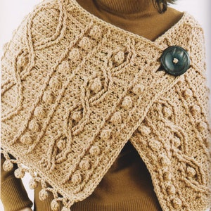 Könnte beinhalten: Ein beigefarbener gehäkelter Schal mit strukturiertem Muster und einem großen, dunklen Knopfverschluss. Der Schal hat ein detailliertes Design mit Noppen und einen Fransenbesatz aus kleinen, dekorativen Kugeln am Rand. Ein gemütliches, handgefertigtes Accessoire.