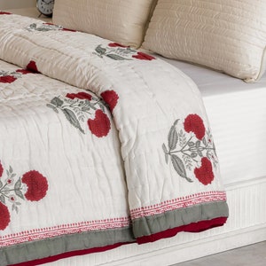 Może przedstawiać: Białe łóżko z białą i czerwoną pikowaną kołdrą. Łóżko ma dwie białe poduszki i dwie kremowe poduszki o subtelnej fakturze. Biała szafka nocna z dwoma szufladami znajduje się po prawej stronie łóżka.