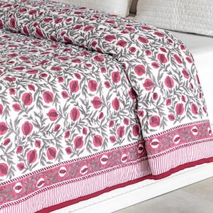 May include: A white and pink floral patterned quilt with grey leaves and stems. The quilt has a border with a striped pattern and a solid pink edge. The bedding is on a white bed frame with white pillows.