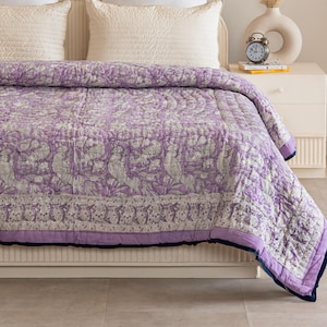 Op de afbeelding: Een paarse en witte quilt met een bloemenpatroon en een donkerblauwe rand. De quilt ligt op een bed met witte kussens en een wit hoofdeinde.